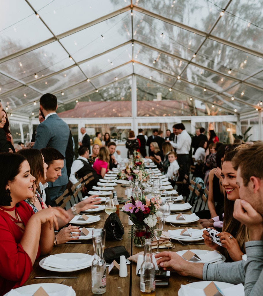 Wedding reception guests under fairy lights in a clear marquee.